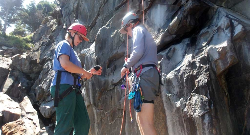 Two people wearing helmets and other safety gear stand near a rock face, preparing to climb. 